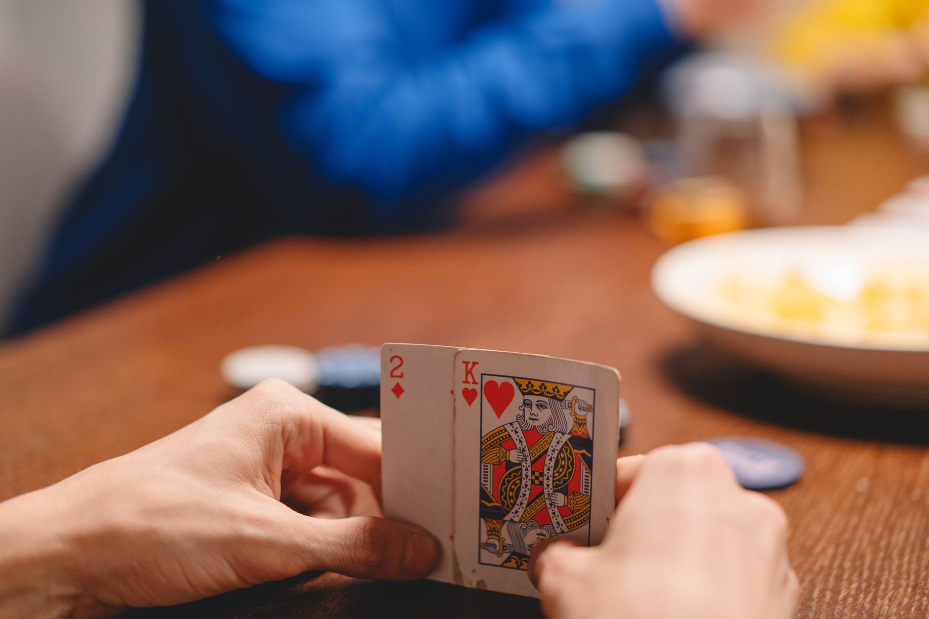 Deux mains tenant un deux de carreau et un roi de cœur sur une table en bois, avec des jetons de poker en arrière-plan flou.